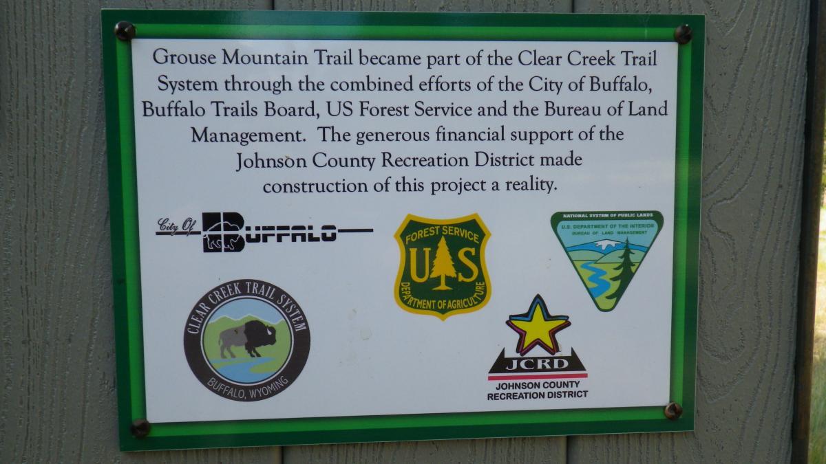 Signage for the Grouse Mountain Trail, part of the Clear Creek Trail System. The sign details the collaboration between the City of Buffalo, Buffalo Trails Board, US Forest Service, Bureau of Land Management, and Johnson County Recreation District to support the trail's development. Various institutional logos are displayed, including those of the City of Buffalo, US Forest Service, and Johnson County Recreation District. Clear Creek mountain bike trail.