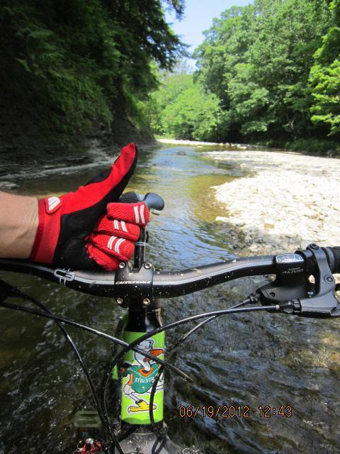 A close-up view of a mountain bike handlebar with a hand wearing a red glove giving a thumbs-up gesture. In the background, a shallow river flows through a lush green landscape, with rocky banks on either side. The image captures a moment of joyful outdoor adventure. Asbury Woods/ Brown's Farm mountain bike trail.