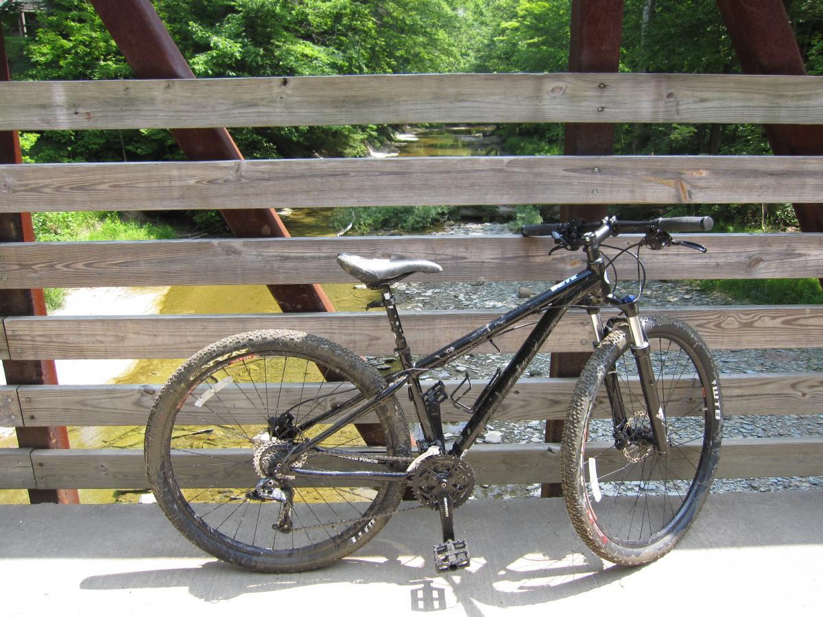A black mountain bike rests on a concrete surface in front of a wooden railing bridge. In the background, a creek flows gently, surrounded by green trees and foliage. The bike shows signs of use, with dirt on the tires. Asbury Woods/ Brown's Farm mountain bike trail.