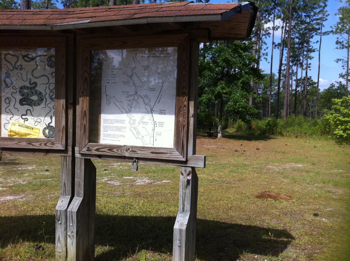 A wooden information kiosk in a grassy area surrounded by tall trees, featuring two panels showing maps and wildlife warnings. One panel includes a detailed map of the trails, while the other displays a cautionary notice. In the background, a picnic table can be seen among the greenery. Bethel Bike Trails mountain bike trail.