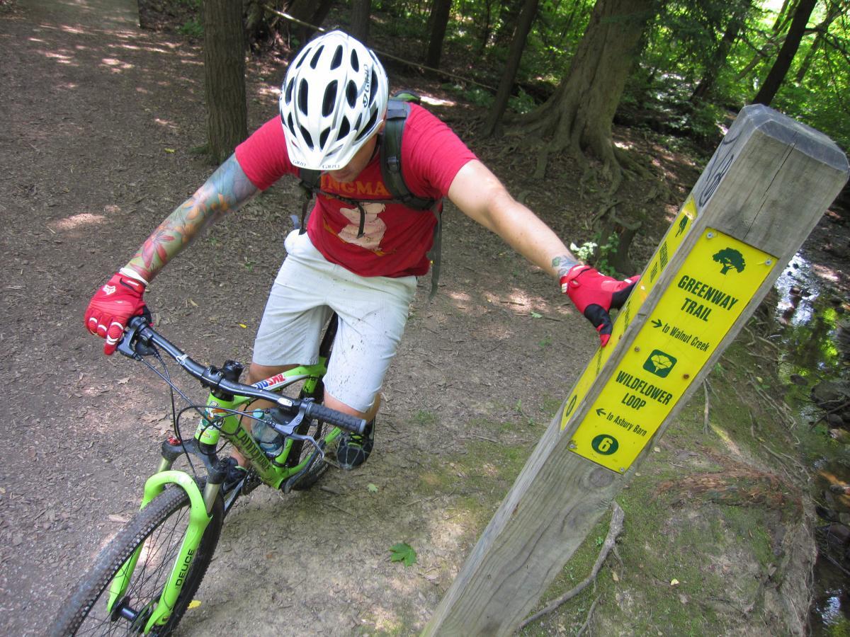 A mountain biker in a red shirt and gloves leans against a green bicycle while examining a trail sign in a wooded area. The sign indicates directions for the Greenway Trail and Wildflower Loop. Sunlight filters through the trees, illuminating the dirt path. Asbury Woods/ Brown's Farm mountain bike trail.