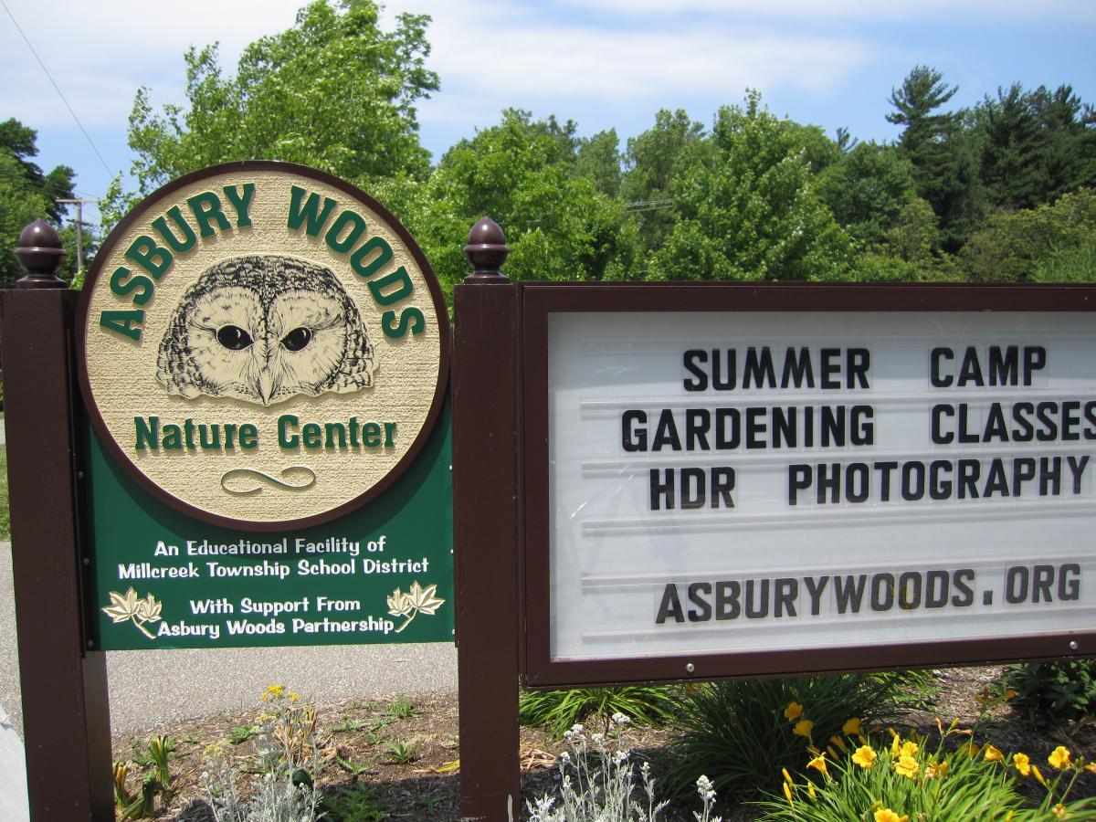 Sign for Asbury Woods Nature Center featuring an owl logo, listing activities such as summer camp, gardening classes, and HDR photography. The sign is surrounded by greenery and flowers, with a clear blue sky in the background. Asbury Woods/ Brown's Farm mountain bike trail.