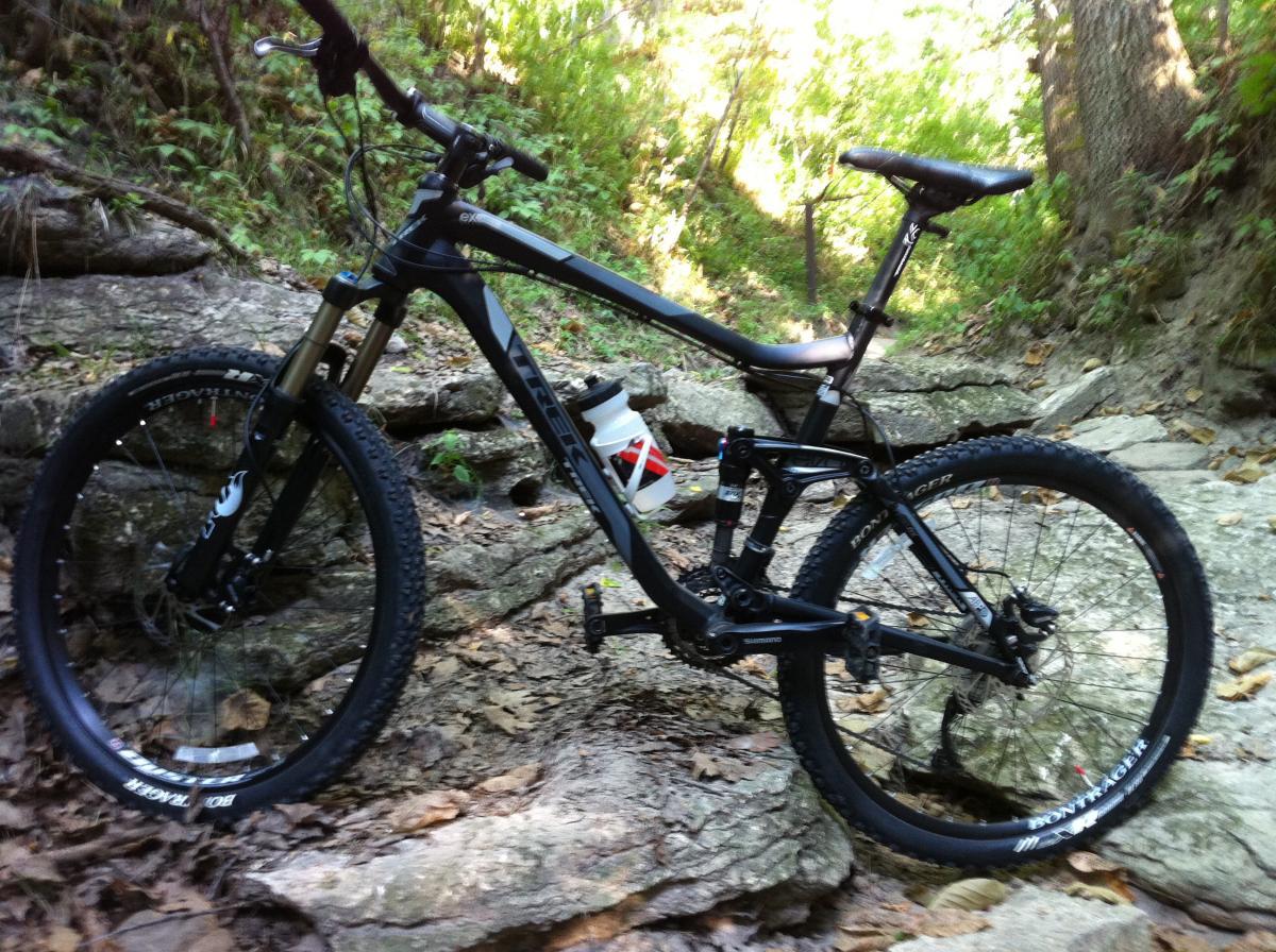 A mountain bike parked on rocky terrain surrounded by greenery, with a water bottle attached to the frame. Platte River mountain bike trail.