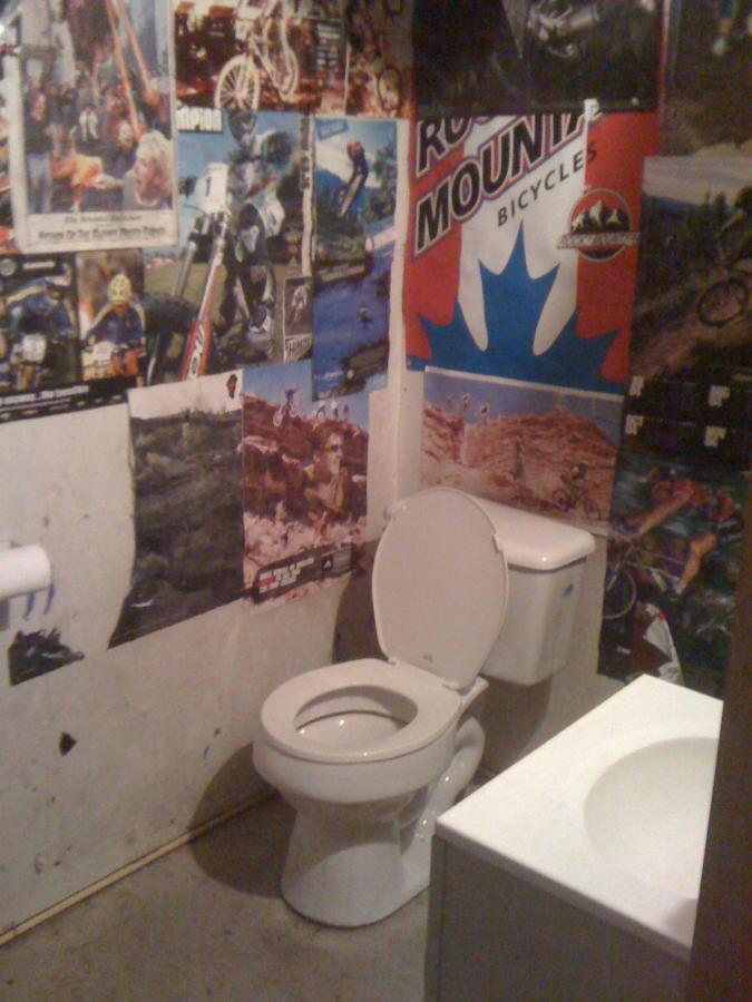 A small bathroom featuring a white toilet with the lid up, adjacent to a white sink. The walls are covered with various colorful posters related to biking and outdoor activities, including a prominent "Rocky Mountain Bicycles" poster. The floor is concrete, and there is a toilet paper holder on the left side of the image. Pee Wee's Mountain Bike Park mountain bike trail.