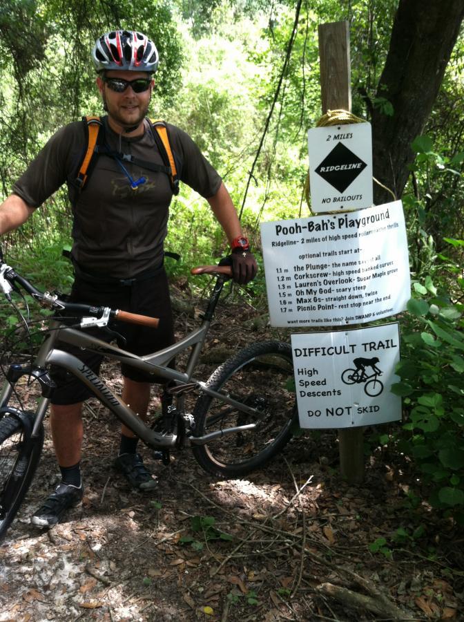A mountain biker stands next to a trail sign marked "Pooh-Bah's Playground," which lists various trails and their difficulty levels. The sign indicates a ridgeline route of 2 miles with high-speed descents and warns not to skid. The biker is wearing a helmet and sunglasses, dressed in a short-sleeved shirt and shorts, with a mountain bike beside him. Lush greenery surrounds the area, indicating a natural outdoor environment. Balm Boyette Scrub Preserve mountain bike trail.