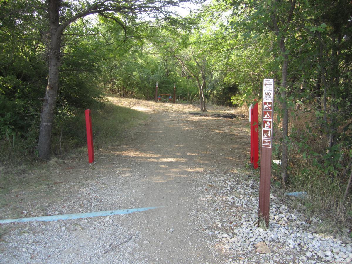 A dirt path surrounded by trees, leading into a wooded area. On either side of the path, red markers are visible, with a signpost displaying various trail rules and restrictions, including a prohibition on motor vehicles. The ground is mostly gravel, and the surroundings are lush and green. Cedar Hill State Park At Joe Pool Lake mountain bike trail.