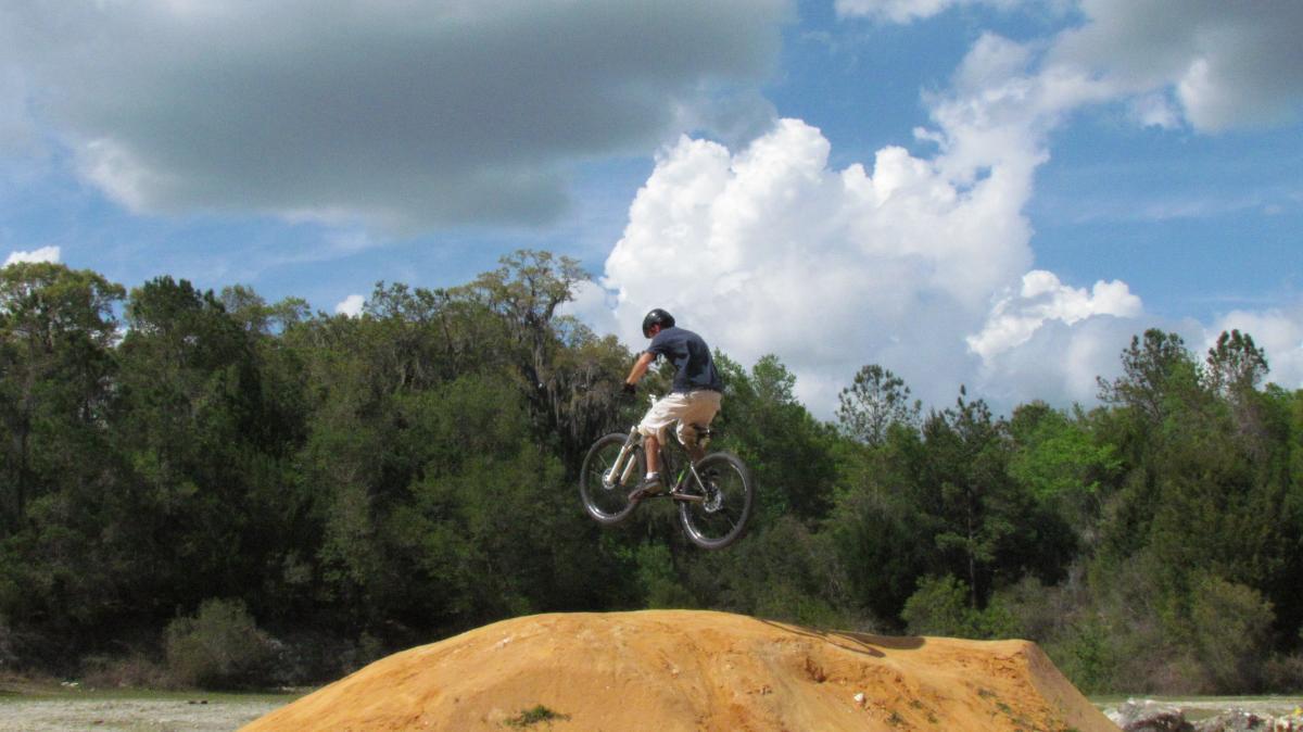 A person in a helmet is performing a jump on a mountain bike above a dirt ramp, surrounded by trees and a blue sky with clouds. Santos mountain bike trail.