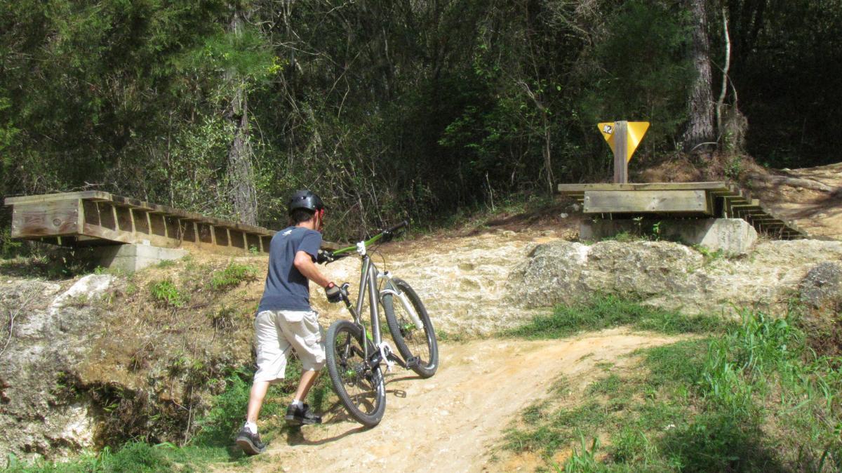A person wearing a helmet is walking up a dirt path while carrying a mountain bike. In the background, there are wooden features, including a ramp and a trail marker, surrounded by trees and greenery. The terrain appears rugged and is designed for biking. Santos mountain bike trail.