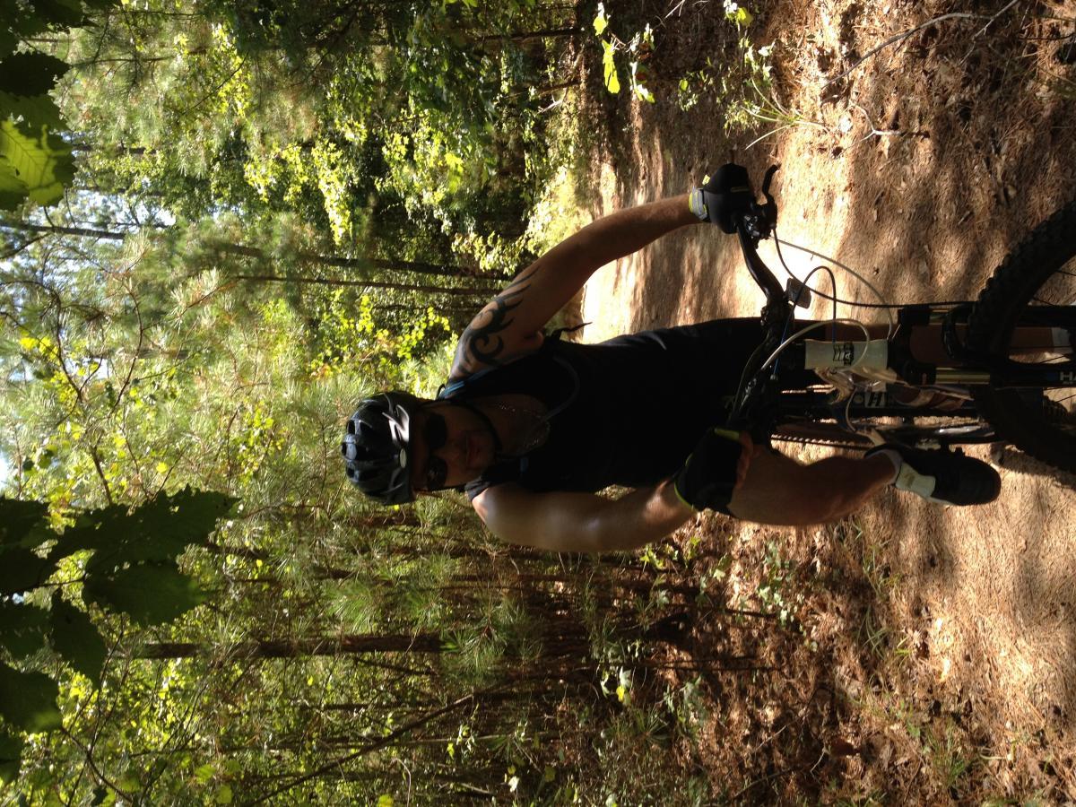 A man in a black tank top and sunglasses is riding a mountain bike on a forested trail. He is wearing a helmet and gloves, surrounded by lush green trees and underbrush, with sunlight filtering through the leaves. Harbison State Forest mountain bike trail.