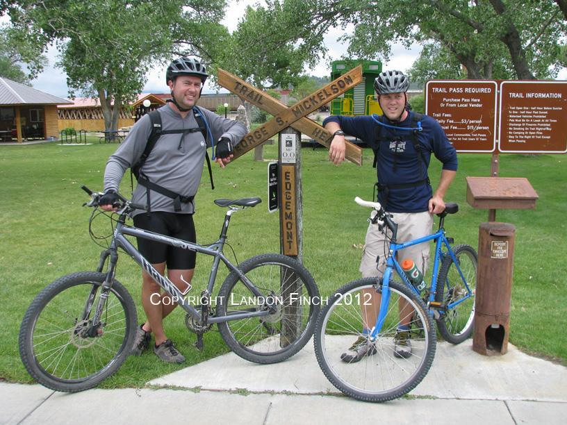 Two men pose next to a wooden signpost indicating the George S. Mickelson Trail and Edgemont, with mountain bikes parked alongside them. The setting features green grass and trees in the background, as well as informational signs about trail passes. One man wears a helmet and a gray athletic shirt, while the other sports a blue shirt and shorts. The George S. Mickelson Trail mountain bike trail.