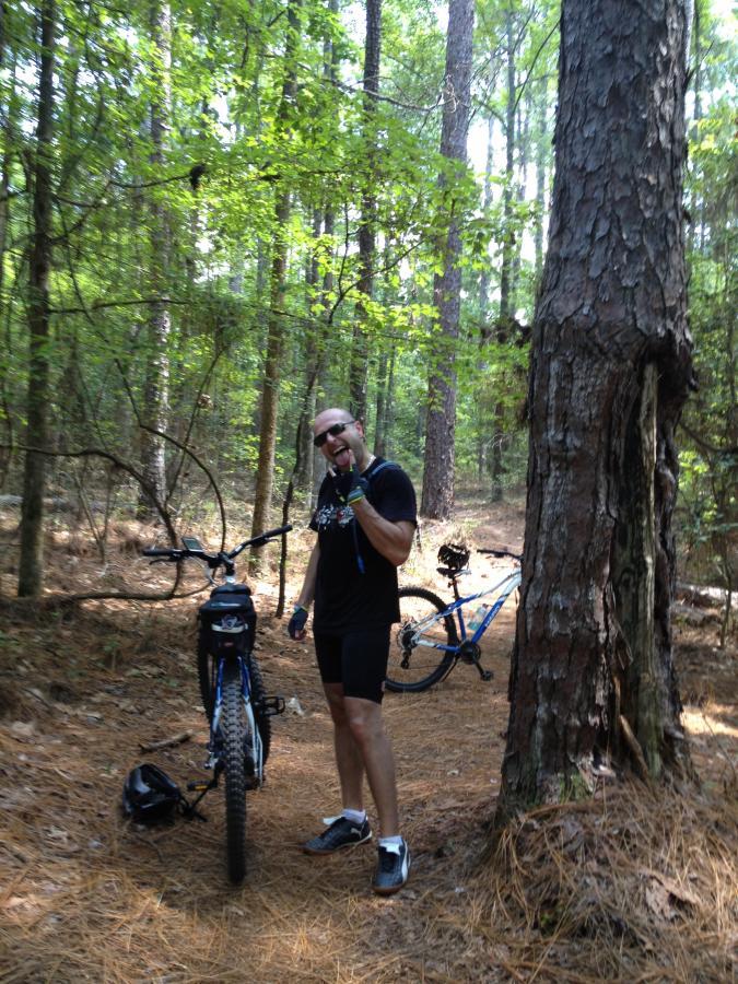 A person in athletic gear stands beside a mountain bike on a wooded trail, smiling and giving a thumbs-up gesture. The background features tall trees and a forest floor covered in pine needles. Another bike can be seen in the distance. Harbison State Forest mountain bike trail.