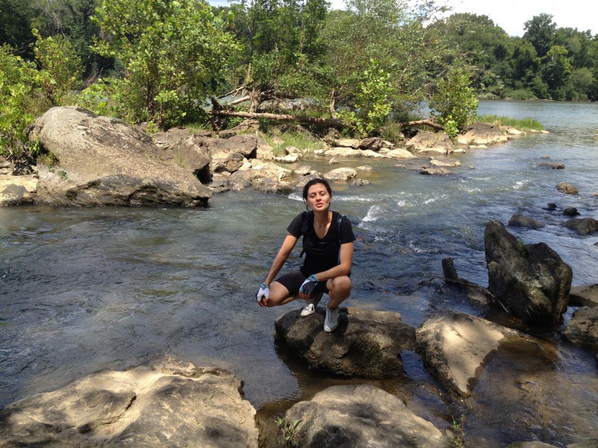 A person crouches on a rock by the edge of a river, surrounded by greenery and smooth stones. The water flows gently in the background, with a mix of sunlight and shade creating a natural setting. The individual is wearing a black t-shirt and shorts, looking relaxed in a serene outdoor environment. Harbison State Forest mountain bike trail.