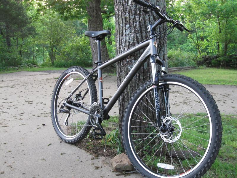 Motobecane 400HT: A mountain bike leaning against a tree in a wooded area, with a visible dirt path and lush greenery in the background.