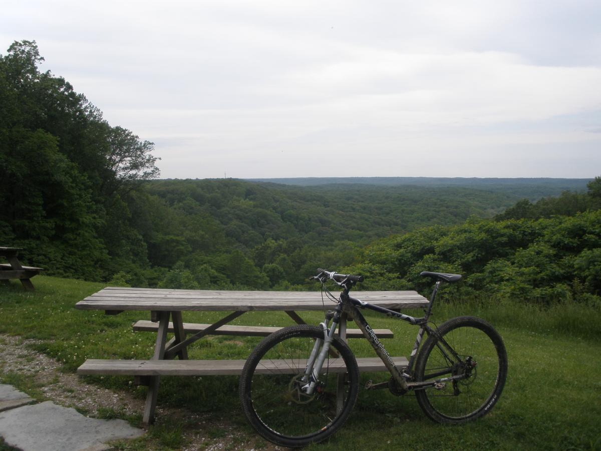 A mountain bike leaning against a picnic table, overlooking a scenic view of lush green hills under a cloudy sky. Brown County Park mountain bike trail.