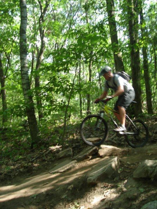 A man riding a mountain bike on a dirt trail through a lush green forest, navigating a rocky section of the path. The image captures the motion and energy of outdoor biking. Russell Mill mountain bike trail.