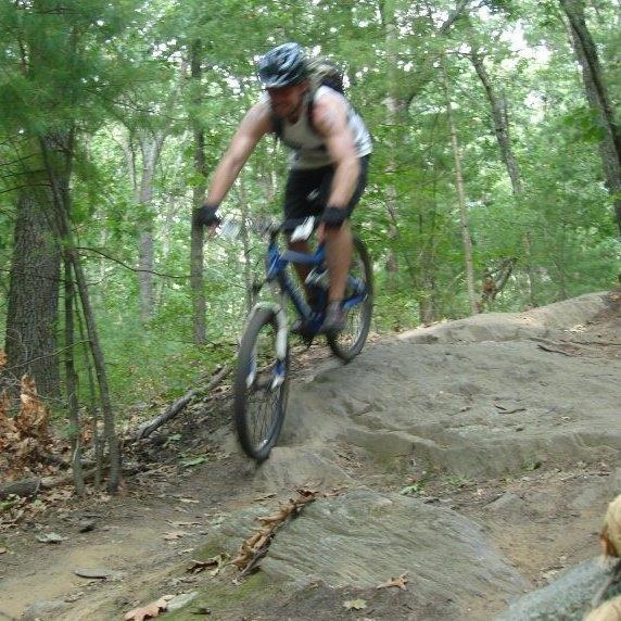 A person riding a mountain bike over a rocky trail in a wooded area. The cyclist is wearing a helmet and a sleeveless shirt, showing a dynamic motion as they navigate the terrain. Russell Mill mountain bike trail.