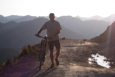 A mountain biker stands on a dirt trail, holding his bike beside him, with a backdrop of rugged mountains under a hazy sky. The sun is low in the sky, casting long shadows and creating a serene atmosphere. A small puddle reflects the surroundings on the trail. Alpine Loop mountain bike trail.