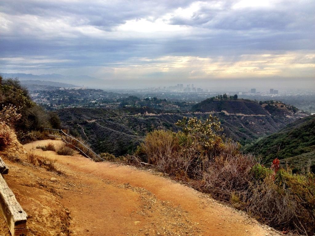 A scenic view from a hiking trail featuring a dirt path winding through hills and vegetation, overlooking a city skyline in the distance under cloudy skies. Backbone Trail: Topanga State Park to Will Rogers State Park mountain bike trail.