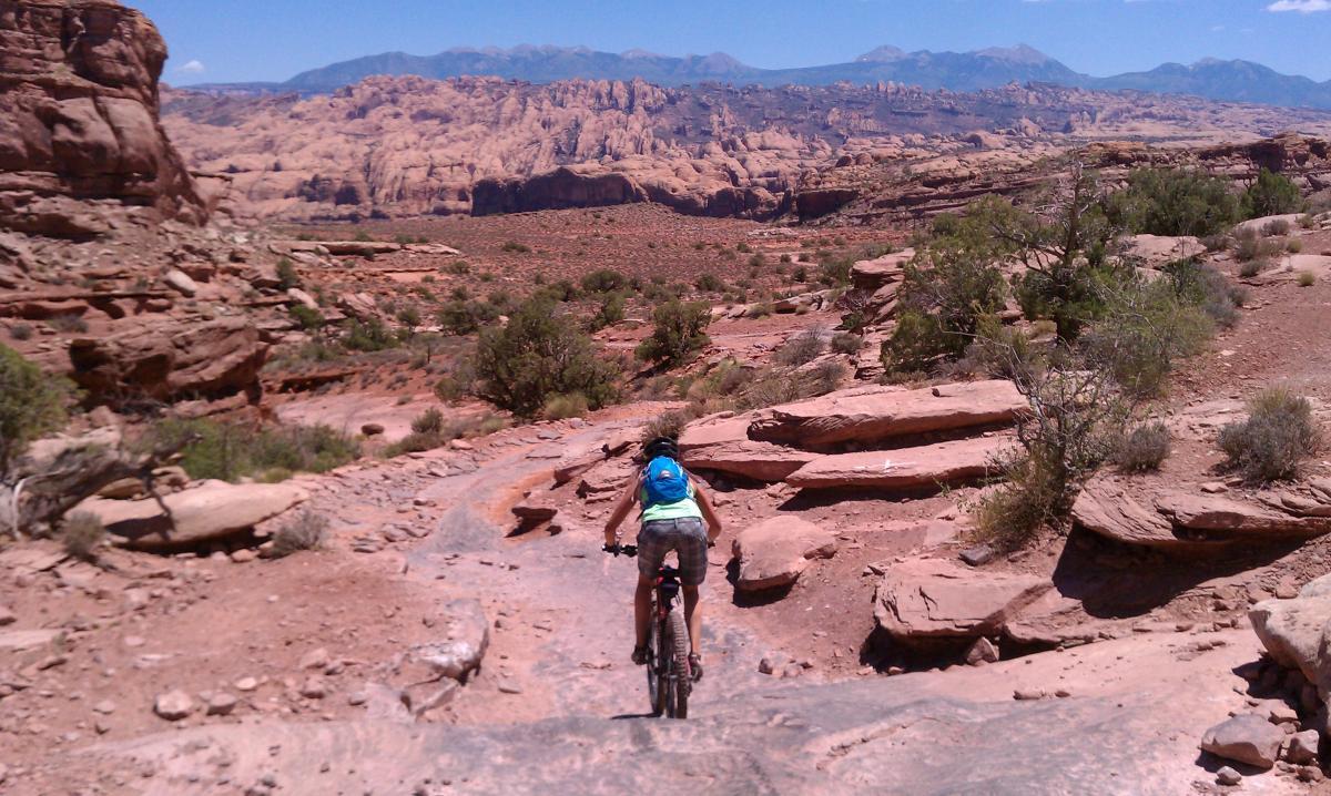 A mountain biker navigates a rocky trail in a desert landscape, surrounded by rugged terrain and distant mountains under a clear blue sky. The scene captures the adventurous spirit of outdoor cycling in a natural setting. Amasa Back Trail mountain bike trail.