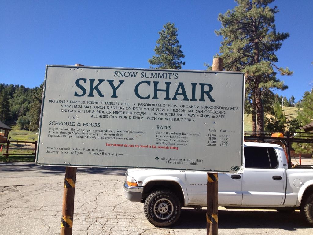 Sign for Snow Summit's Sky Chair showing details about the scenic chairlift ride. Features include a panoramic view of the lake and surrounding mountains, along with information about hours of operation, rates for adult and child rides, and guidelines for biking. In the background, there is a truck parked near the sign and trees lining the area. Big Bear Mountain Resort mountain bike trail.