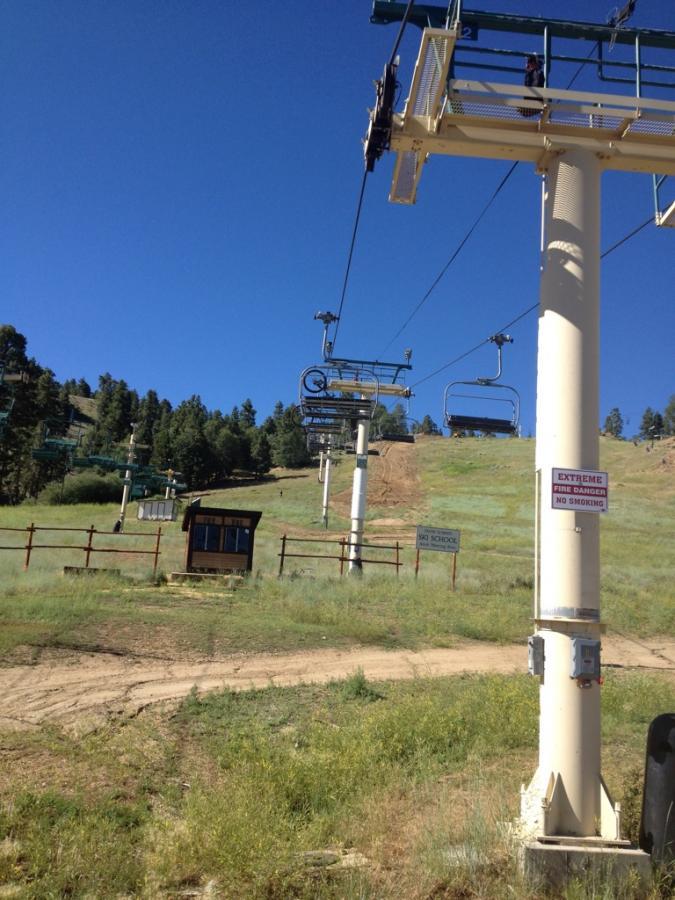 A ski lift with empty chairs ascends a green hillside under a clear blue sky. In the foreground, a wooden ticket booth is visible beside a sign that reads "No School" and another sign warning about "Extreme Fire Danger" and "No Smoking." Tall trees are scattered along the slope. Big Bear Mountain Resort mountain bike trail.