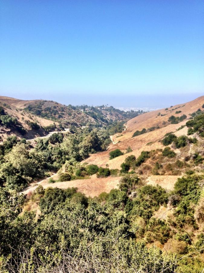 A panoramic view of rolling hills and valleys with sparse vegetation under a clear blue sky. The landscape features a mix of brown and green areas, with a winding path visible in the lower section, suggesting a natural and rugged terrain. The distant horizon shows a slight haze, typical of warm days. Turnbull Canyon Trail mountain bike trail.