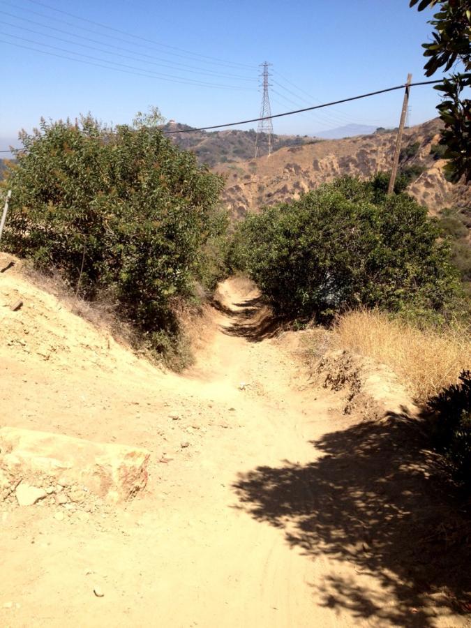 A dirt trail winding through a hillside, surrounded by shrubs and grasses, under a clear blue sky. Power lines run across the background, with rocky terrain visible on both sides of the path. Turnbull Canyon mountain bike trail.
