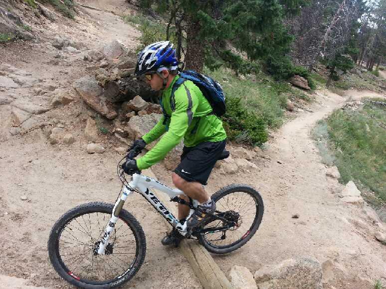 A mountain biker in a bright green jacket and helmet navigates a rocky trail on a white bicycle, focused on the path ahead. The surrounding area features trees and dirt paths, indicating a rugged outdoor environment. 3 Sisters / Alderfer mountain bike trail.