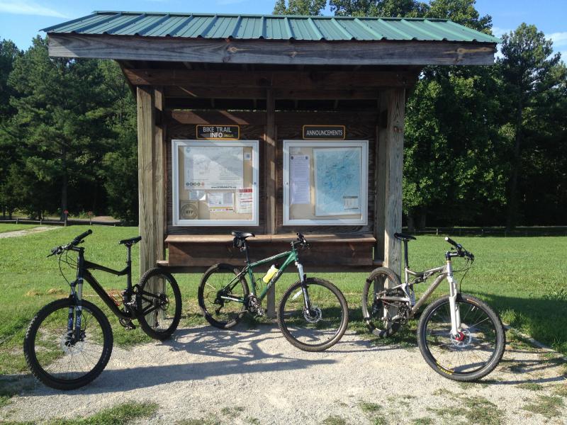 A wooden kiosk with a green roof displaying bike trail information and announcements. Four mountain bikes are parked in front on a gravel path, with a grassy area and trees in the background. Montgomery Bell State Park Mtb Trail mountain bike trail.