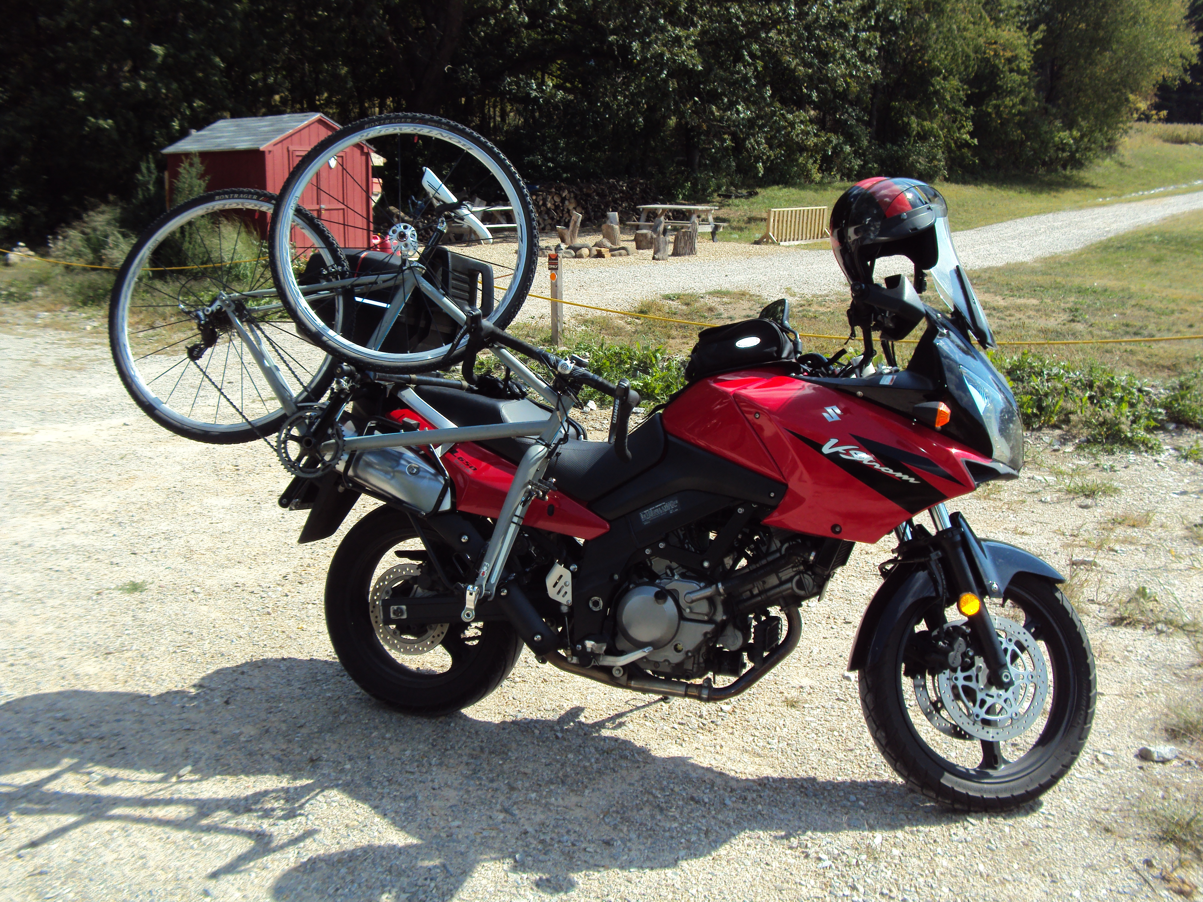 A red motorcycle parked on a gravel path, with a bicycle mounted on its rear rack. The background features a wooded area and a small shed, with outdoor seating visible in the distance.