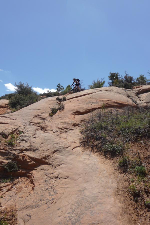 A person hiking on a rocky incline under a clear blue sky, surrounded by sparse vegetation and shrubs. Porcupine Rim mountain bike trail.
