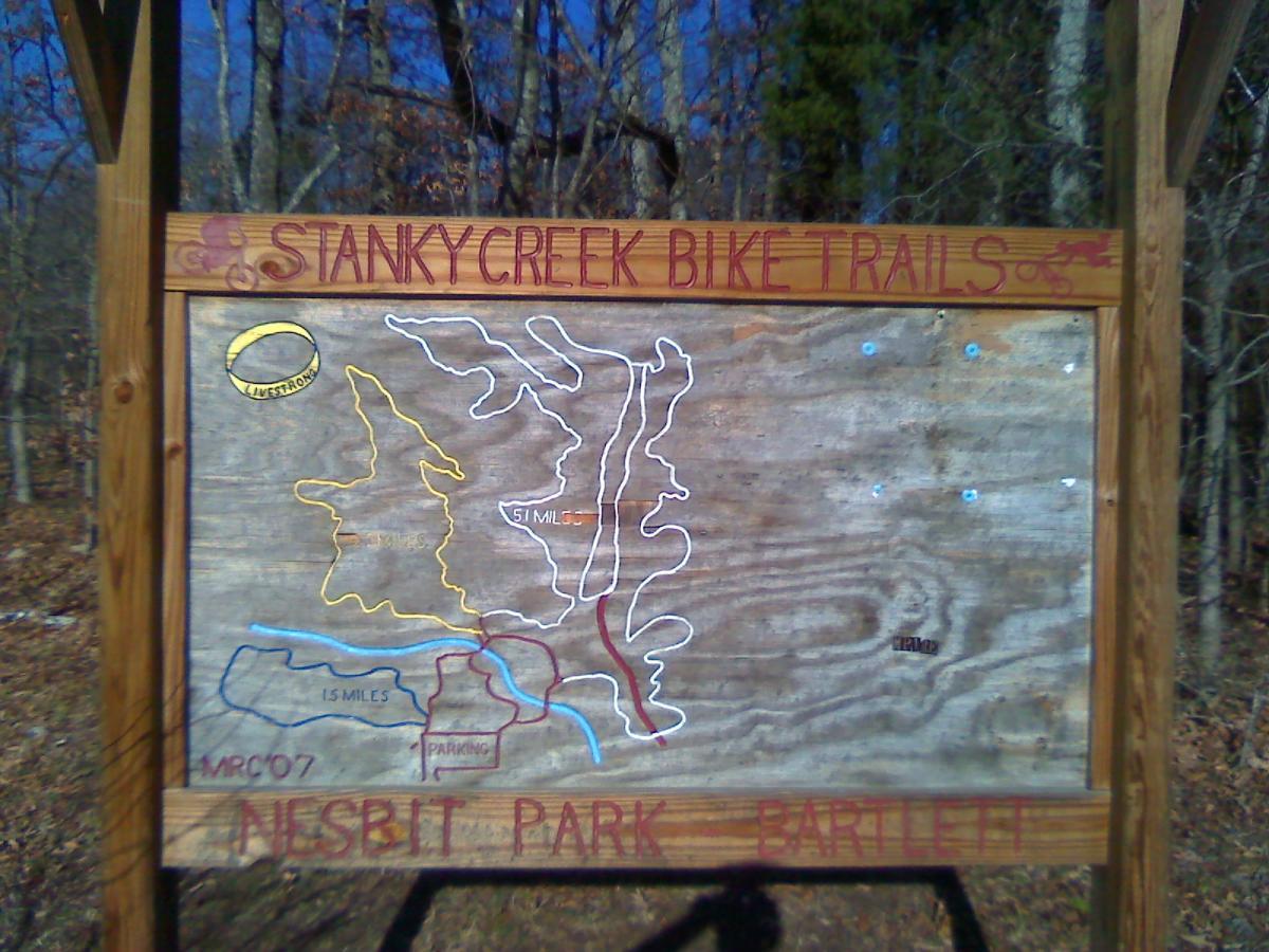 A wooden trail map sign for Stanky Creek Bike Trails, located in Nesbit Park, Bartlett. The sign features a detailed map outlining various biking routes marked in different colors, indicating trail lengths and parking areas. The background shows a wooded area, suggesting an outdoor recreational setting. Stanky Creek mountain bike trail.