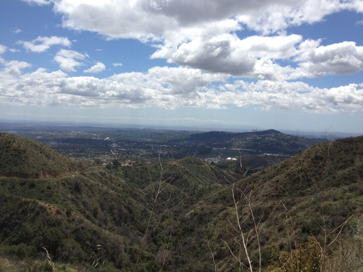 A panoramic view of a hilly landscape under a partly cloudy sky. The foreground features rolling green hills, while the background showcases a distant urban area with a skyline visible. The overall scene conveys a sense of natural beauty and open space. El Prieto mountain bike trail.