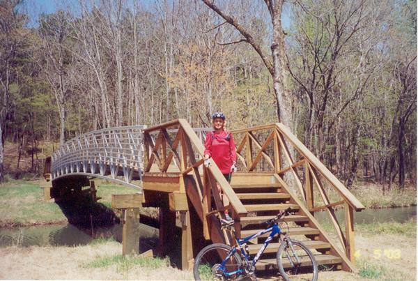 A person in a red shirt and helmet stands next to a wooden bridge and staircase beside a creek. A blue bicycle is positioned at the bottom of the stairs. The scene is set in a wooded area with trees showing early signs of spring foliage. Cedar Lake Trail mountain bike trail.