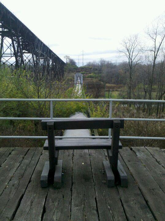 A wooden bench sits on a weathered wooden platform, facing a narrow pathway that leads to a distant bridge surrounded by greenery and trees. In the background, a large metal structure spans overhead, adding a stark contrast to the natural surroundings under a cloudy sky. Ohio And Erie Canal mountain bike trail.