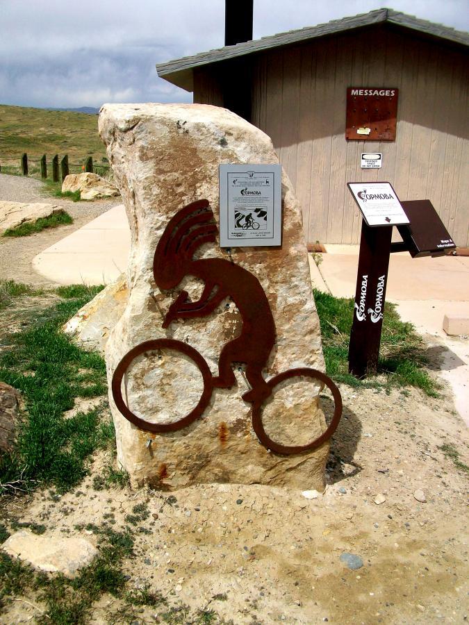 A large rock with a metal silhouette of a cyclist, depicting a person riding a bicycle. Nearby, there are informational signs and a structure in the background labeled "Messages." The setting features grassy terrain under a cloudy sky. Kokopelli Trail mountain bike trail.