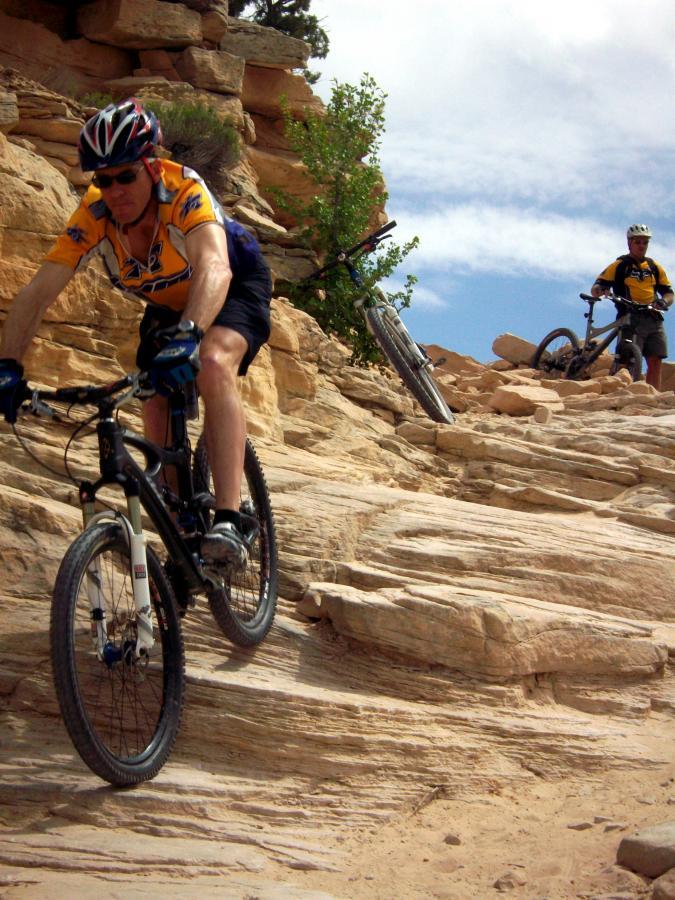 Two mountain bikers navigate a rocky trail, with one rider actively descending a steep, uneven surface. The cyclist in the foreground is wearing a bright yellow jersey and a helmet, focused on maintaining balance. In the background, another cyclist is paused on the trail, with their bike resting against a rocky outcrop. The scene is set against a backdrop of rugged terrain and blue skies. Mary's Loop / Horsethief Bench mountain bike trail.