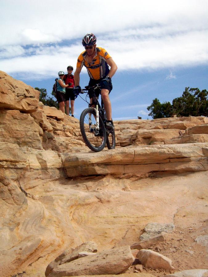 A mountain biker navigates a rocky terrain with steep slopes, while two people in the background observe the ride under a blue sky with scattered clouds. Mary's Loop / Horsethief Bench mountain bike trail.
