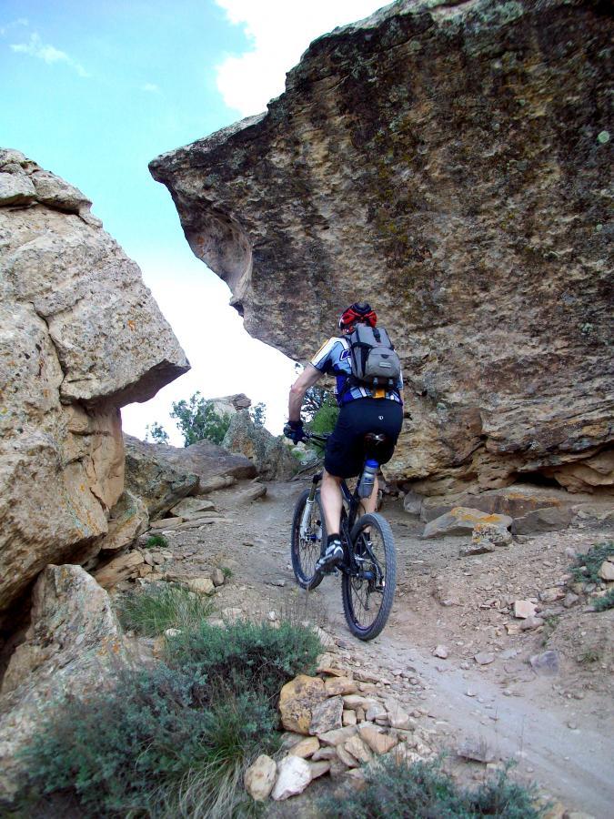 A mountain biker navigates a rocky trail surrounded by large boulders under a blue sky. The path is uneven and dusty, with patches of grass and small shrubs alongside the trail. Mary's Loop / Horsethief Bench mountain bike trail.