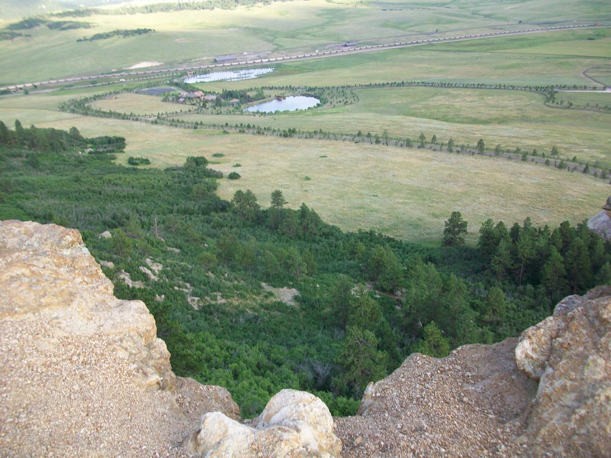 A panoramic view from a rocky cliff overlooking a lush green valley. The landscape features a large, winding pond surrounded by trees, with open fields and distant hills in the background. A line of structures is visible along the horizon, indicating human presence in the natural setting. Spruce Mountain Trail Upper Loop mountain bike trail.