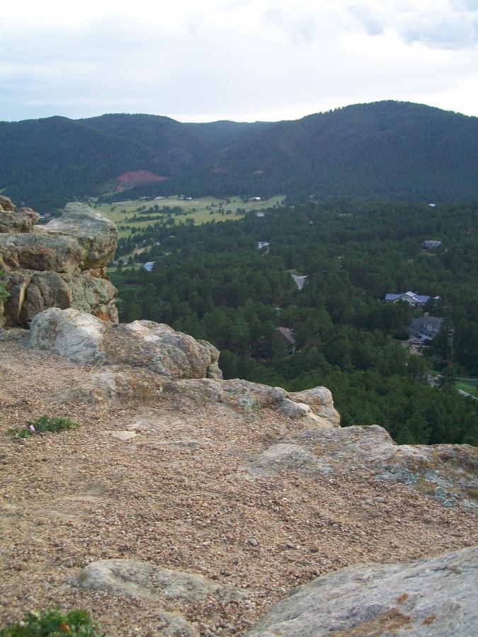 A scenic view from a rocky outcrop overlooking a forested valley with scattered houses. In the background, rolling hills are visible under a cloudy sky. The foreground features rocky terrain with some greenery. Spruce Mountain Trail Upper Loop mountain bike trail.