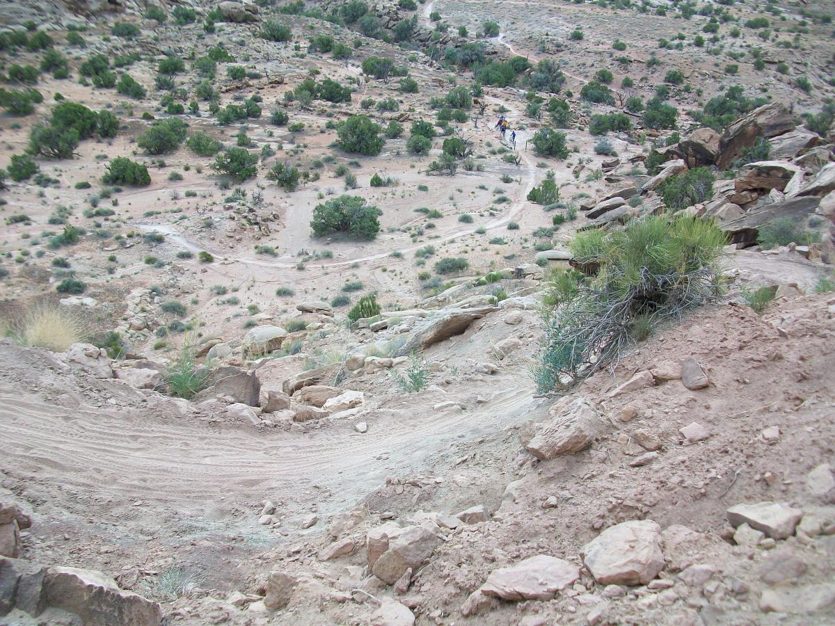 A rocky hillside with a dirt path winding down through sparse vegetation, including small shrubs and bushes, leading into a rugged landscape. In the distance, a couple of figures are seen along the trail. The terrain appears dry and arid, typical of a desert environment. Sovereign Single Track mountain bike trail.