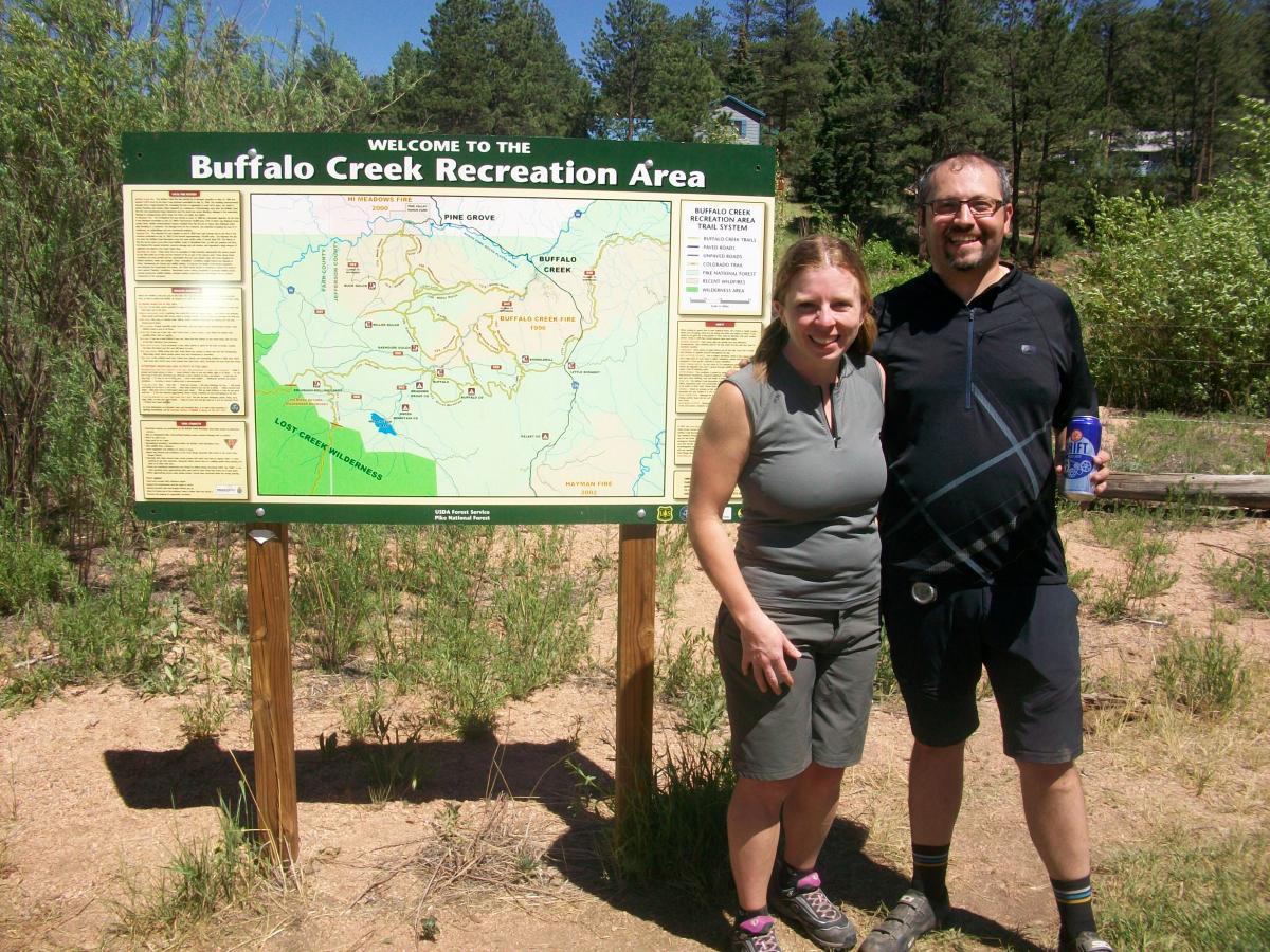 A man and a woman stand in front of a welcome sign for the Buffalo Creek Recreation Area, which displays a map and information about the area. They appear to be enjoying a sunny day outdoors, with greenery and trees in the background. The woman is wearing a sleeveless top and the man is holding a drink. Buffalo Creek mountain bike trail.