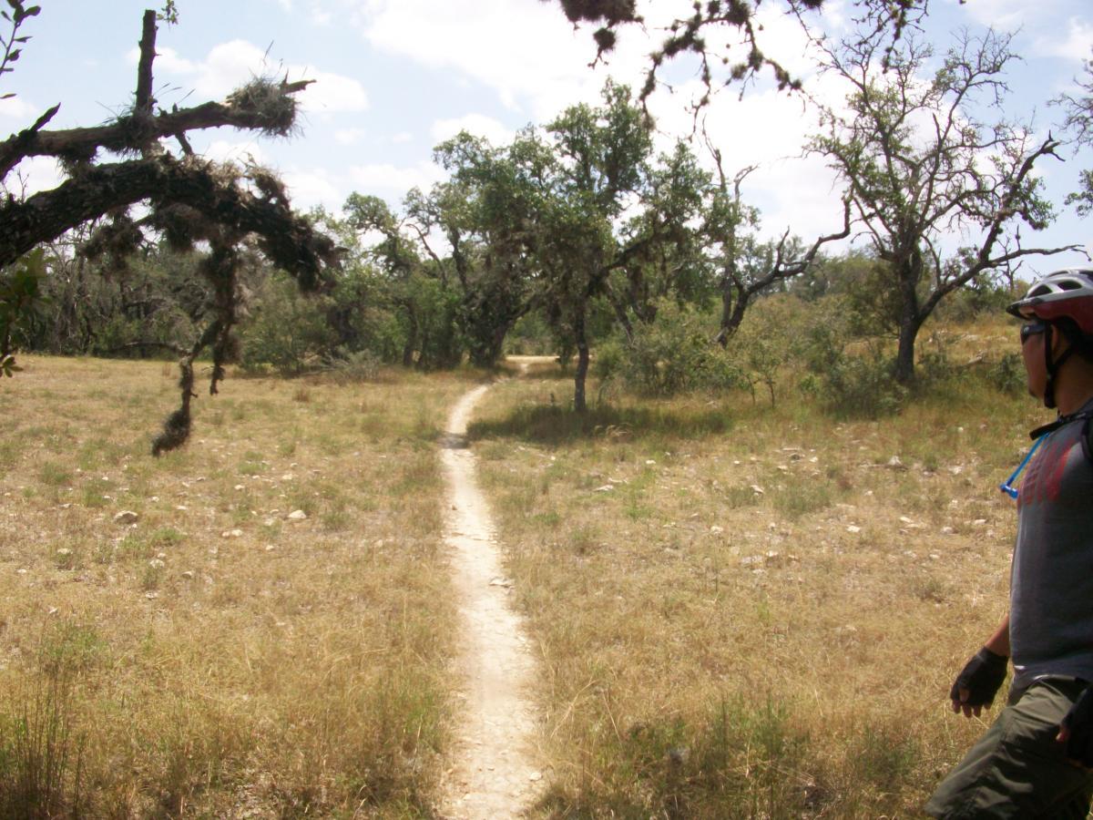 A cyclist stands on a dirt path in a grassy area surrounded by trees. The landscape features sparse vegetation and a clear blue sky with scattered clouds. The cyclist is wearing a helmet and a short-sleeved shirt, looking toward the trail ahead. Flat Rock Ranch mountain bike trail.