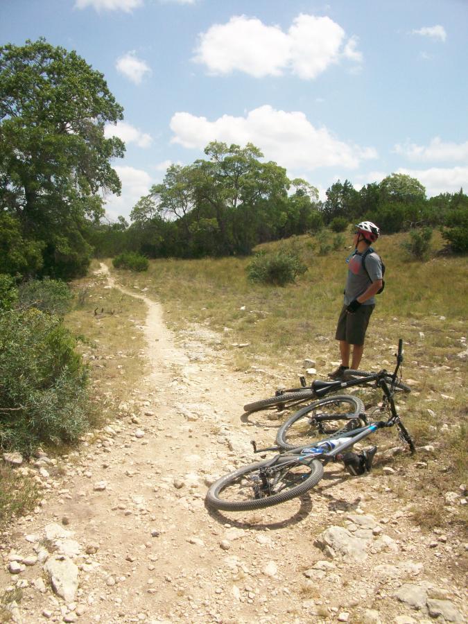 A person in a helmet stands beside two mountain bikes lying on a rocky, dirt path. The scene is set in a grassy area with scattered trees under a partly cloudy sky, suggesting a warm and sunny day. The individual appears to be looking thoughtfully at the trail ahead, indicating a moment of pause during a biking adventure. Flat Rock Ranch mountain bike trail.