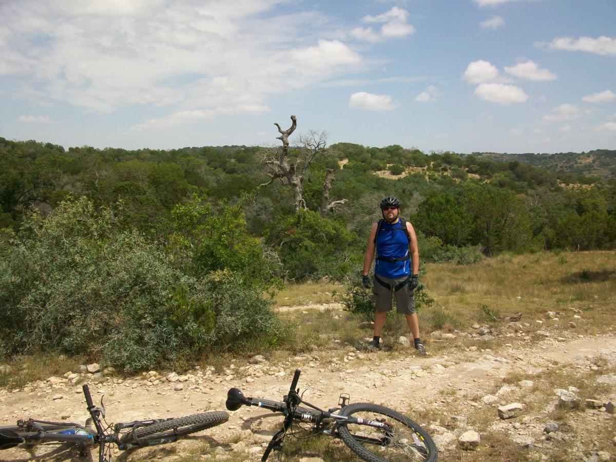 A person stands on a dirt trail surrounded by lush greenery and hills, wearing a blue tank top and a bike helmet. Two mountain bikes lie on the ground in the foreground, partially obscured by grass. The sky is bright with scattered clouds, creating a vibrant outdoor scene. Flat Rock Ranch mountain bike trail.