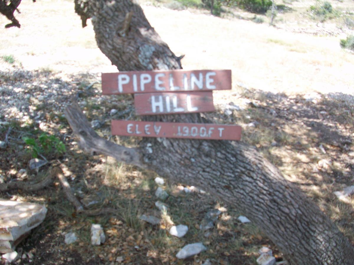 Sign marking "Pipeline Hill" at an elevation of 1900 feet, mounted on a tree trunk, surrounded by rocky ground and sparse vegetation. Flat Rock Ranch mountain bike trail.