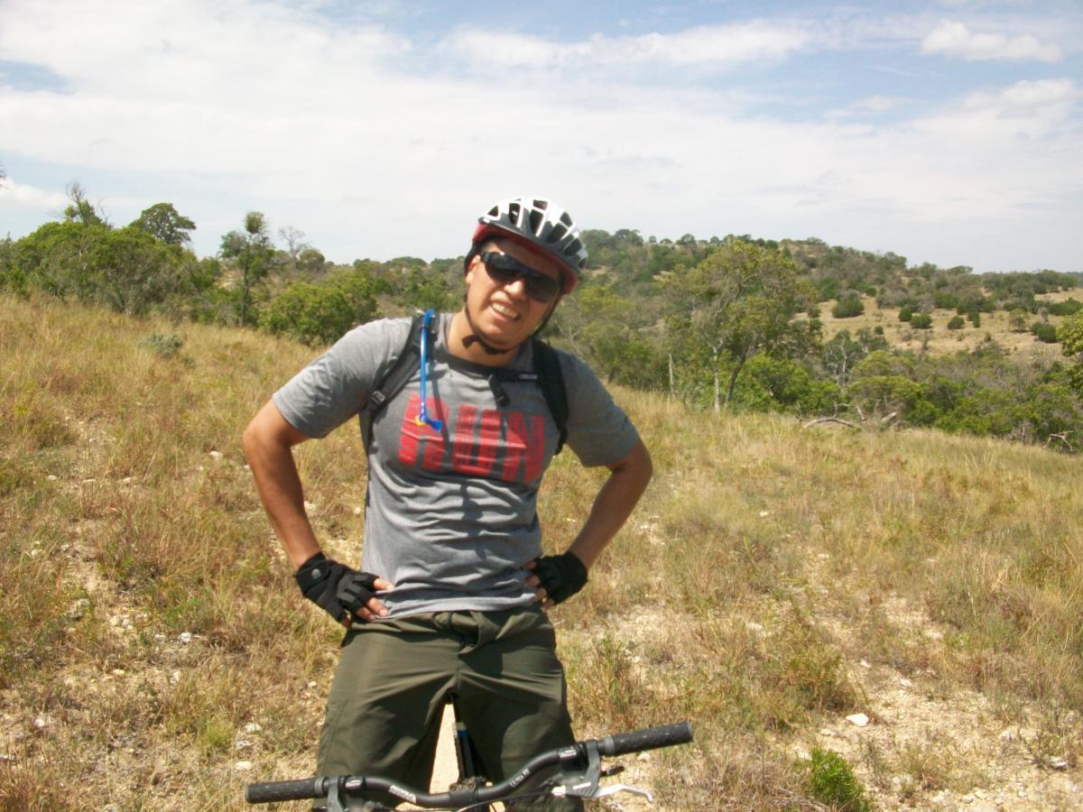 A young man standing next to a mountain bike in a grassy landscape, wearing a helmet and sunglasses. He has a casual, sporty outfit with a graphic t-shirt and shorts. The background features rolling hills and scattered trees under a blue sky with a few clouds. Flat Rock Ranch mountain bike trail.
