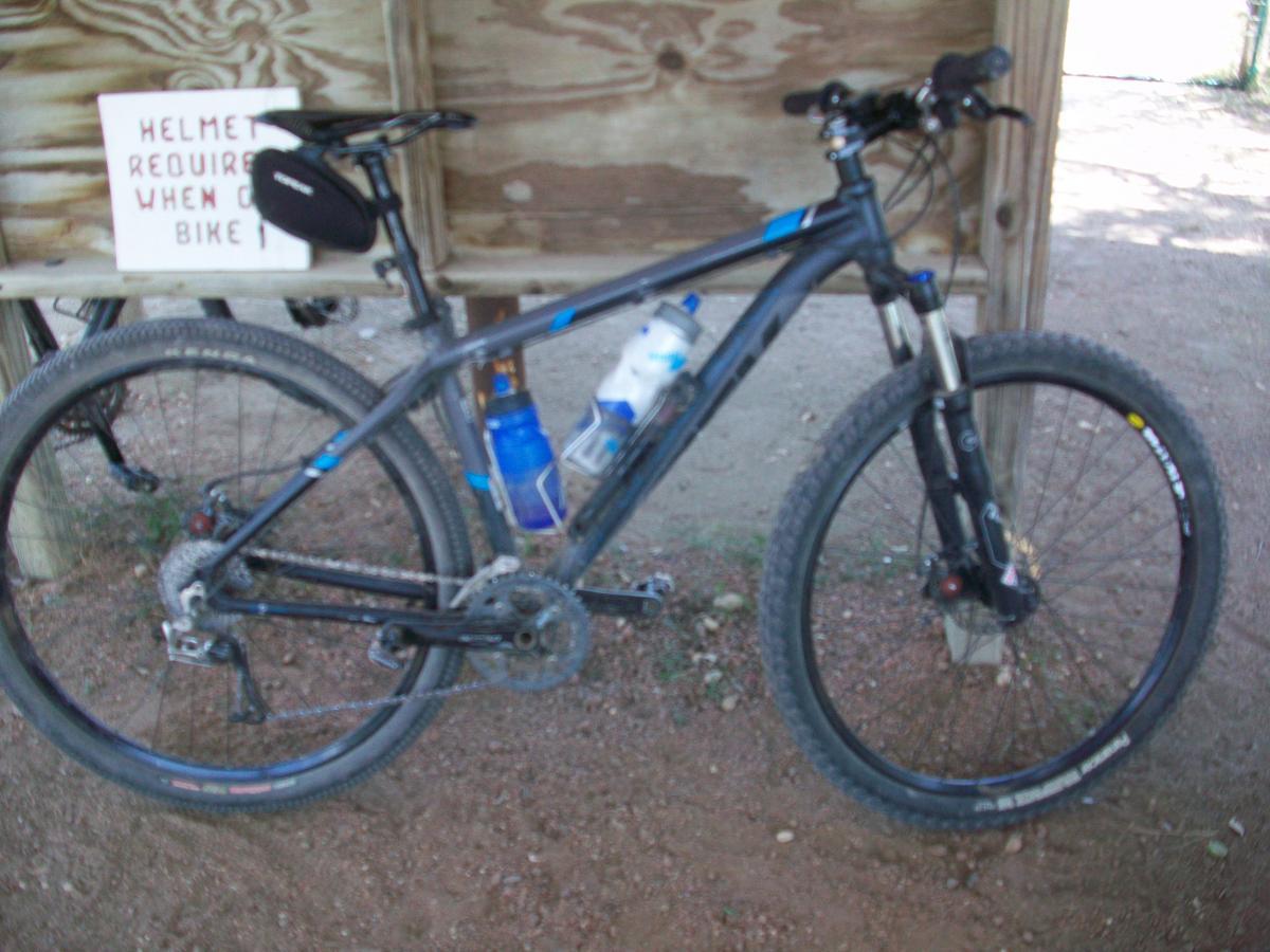 A black mountain bike with blue accents parked next to a wooden sign that reads "Helmet required when biking." Two water bottles are mounted on the bike frame. The background features a dirt path and a wooden structure. Flat Rock Ranch mountain bike trail.