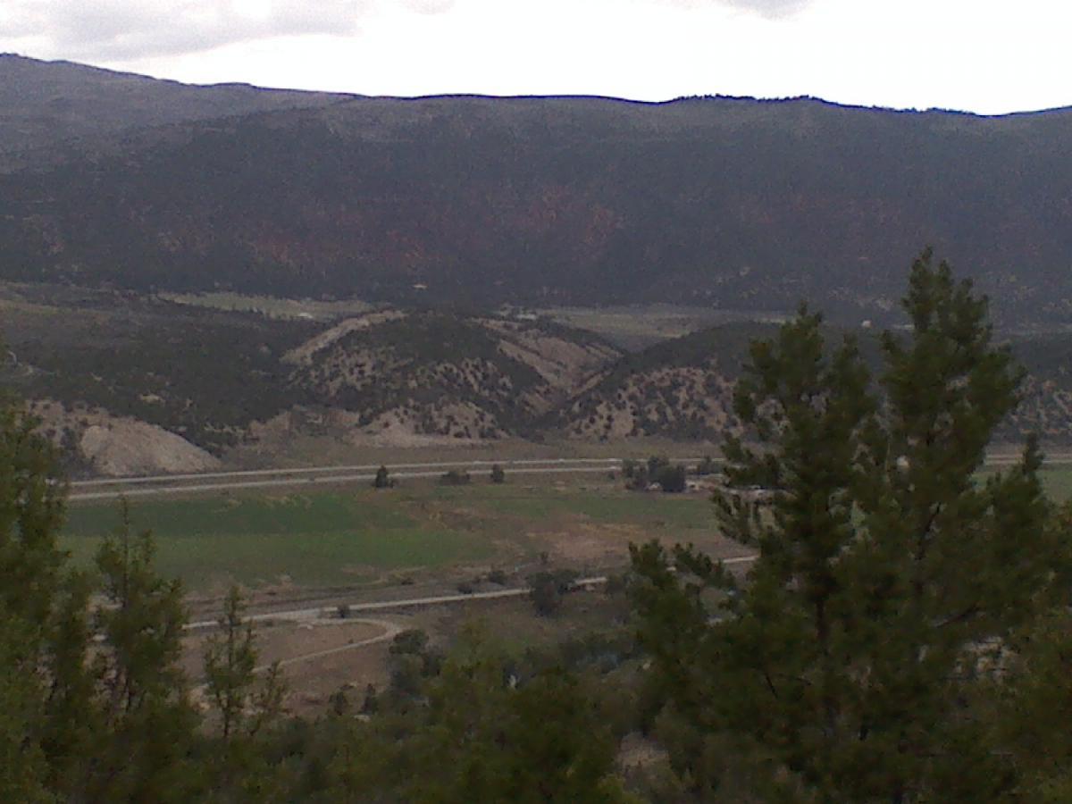 A scenic view of rolling hills and mountains under a cloudy sky, featuring green fields and a meandering road in the foreground, framed by trees on the left side. The Boneyard mountain bike trail.